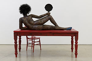 Alison Saar / 
Set to Simmer, 2019 / 
wood, ceiling tin, enamel paint ceiling tin wire, found table, chair, and skillet with texts by Dionne Brand / 
Table and figure: 65 x 72 x 36 in. (165.1 x 182.9 x 91.4 cm) / 
Chair: 38 x 16 x 16 in. (96.5 x 40.6 x 40.6 cm)